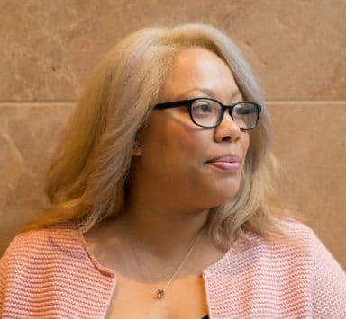 A Black lesbian woman stands in front of a stone wall in an upscale office building. She's wearing a pink cardigan, and necklace with a pink stone. She has blonde hair and wears glasses, and has a confident expression on her face.