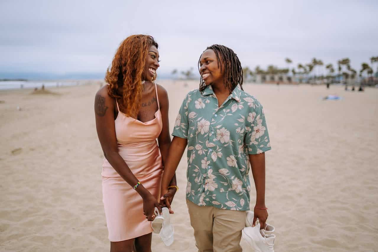 A lesbian couple walks on the beach, smiling hand in hand. The partner on left is a Black femme with long hair in a pink slip dress. The partner on the right is a Black butch woman wearing a floral print button down and braids.
